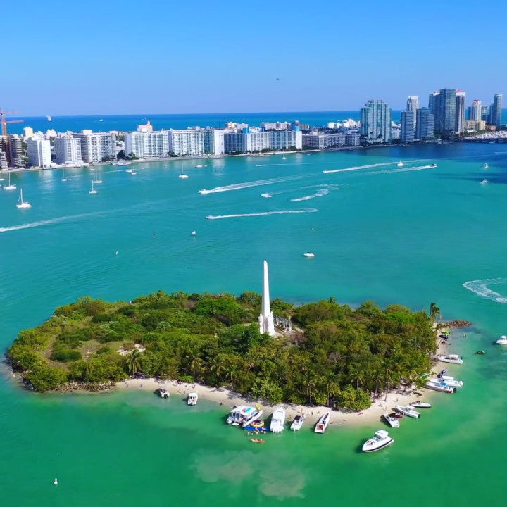 Aerial view the monument island surrounded by turquoise waters and a city skyline in the background.